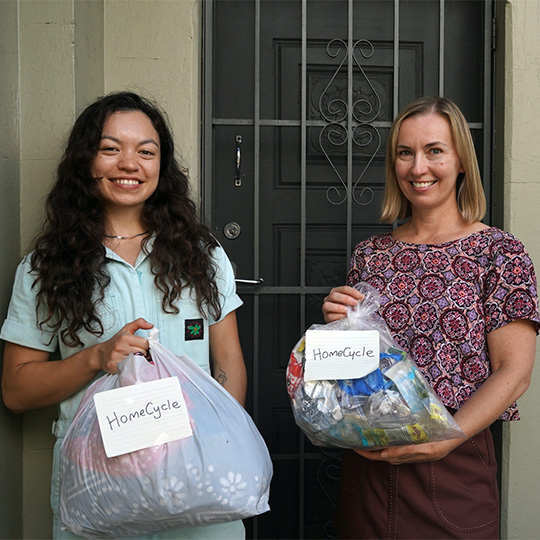 Two people standing by the front door of a house, each holding a bag labeled 'HomeCycle'.